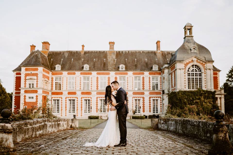 couple homme et femme en tenue de mariage, lui en costume elle en robe de mariée, devant un chateau prestigieux, lieu de réception grandiose