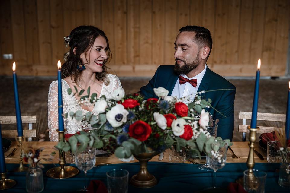 couple de mariés, elle en robe blanche lui en costume bleu, assis derrière la table d'honneur aux couleurs rouge, bleue et blanc, deco de mariage d'hiver rouge et bleue moderne