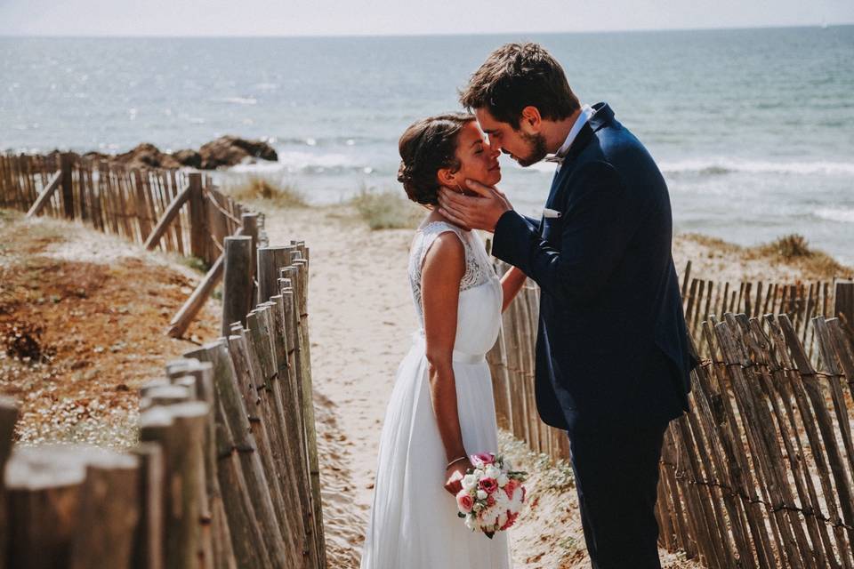 couple de mariés homme et femme en tenue de mariage, elle en robe de mariée blanche et lui en costume bleu marine, sur le point de s'embrasser dans un décor magnifique sur la plage, mariage en bord de mer