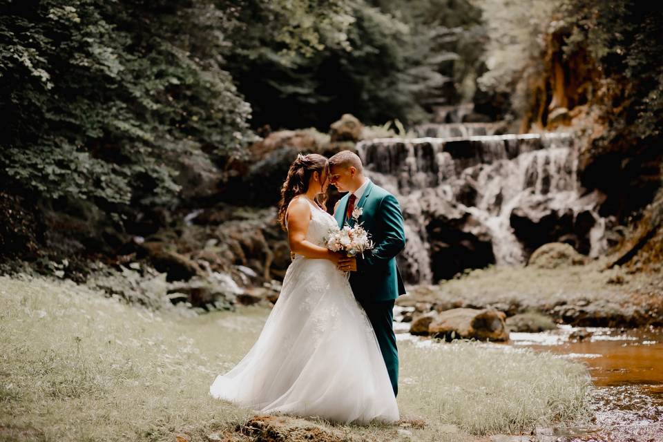 Couple de marié près d'une cascade, la mariée porte une robe princesse et le marié un costume vert