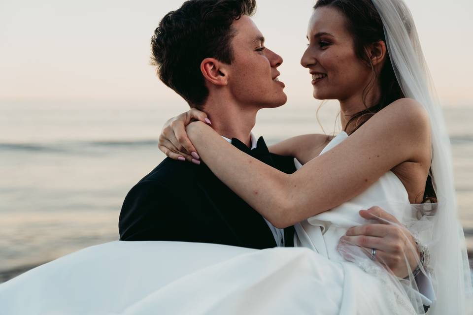 Le marié vêtu d'un smoking porte la mariée dans ses bras qui a une longue robe blanche et un voile, en toile de fond la mer