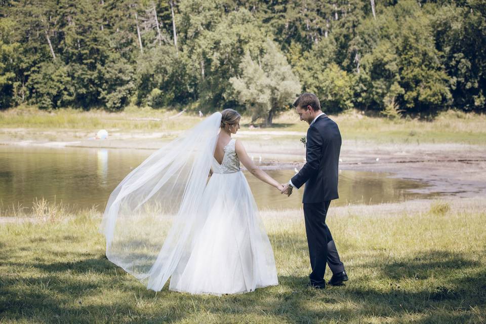 Jeune couple de marié près d'un lac, la mariée porte une robe coupe princesse avec un long voile et son mari un smocking