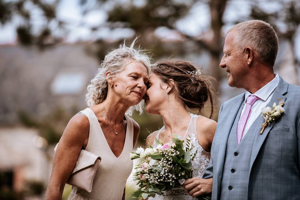 Mariée avec ses deux parents à ses côtés avec un bouquet de fleur rose à la main et des fleurs dans les cheveux son père à une fleur assorti sur sa veste 