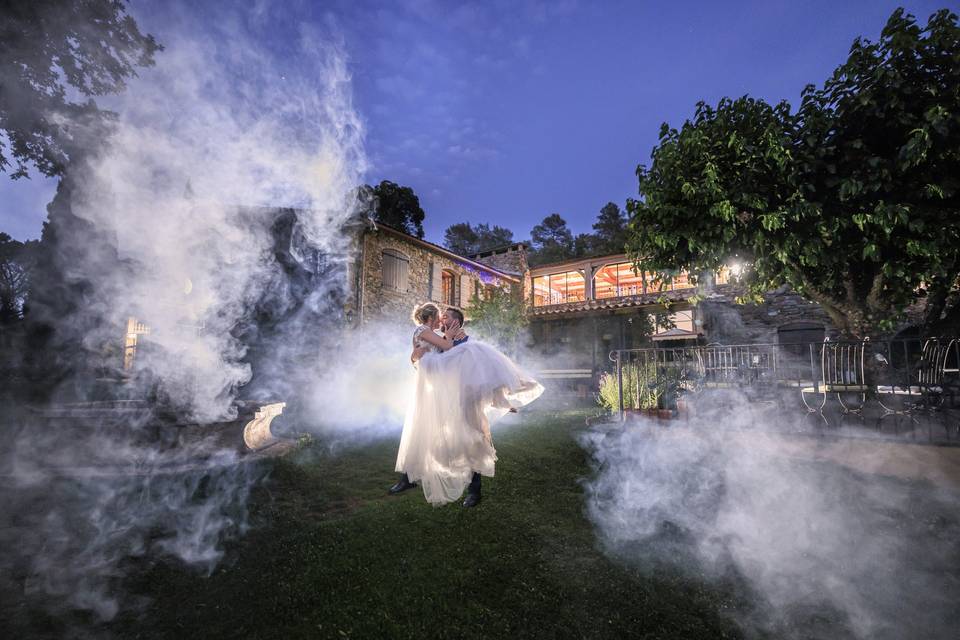 Couple qui s'embrasse, le marié porte la mariée qui a une robe blanche princesse il y a des jet de fumé artificielle autour d'eux, dans un domaine