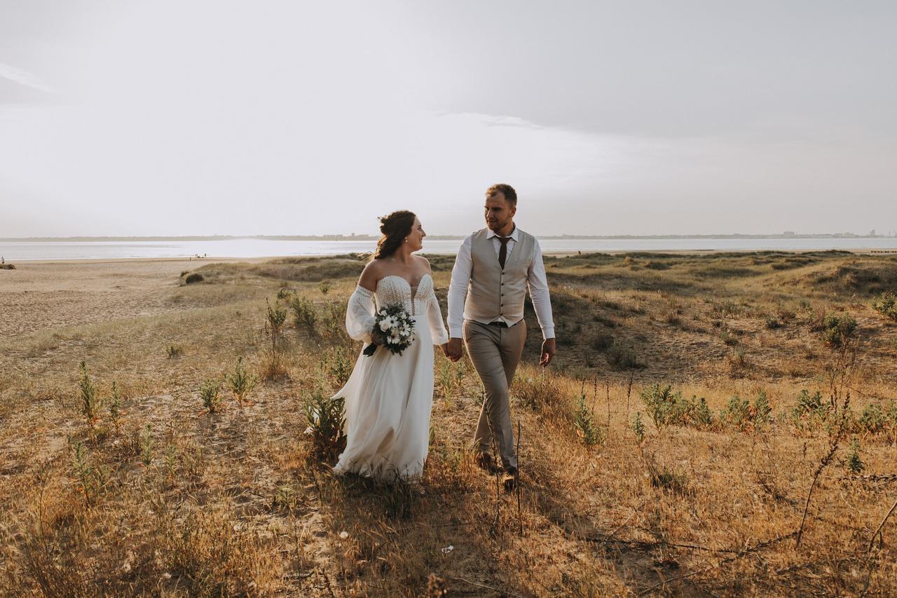 Couple de mariés près de la mer, la mariée a une robe corset évasée et le marié à un ensemble gris avec un gilet de serveur