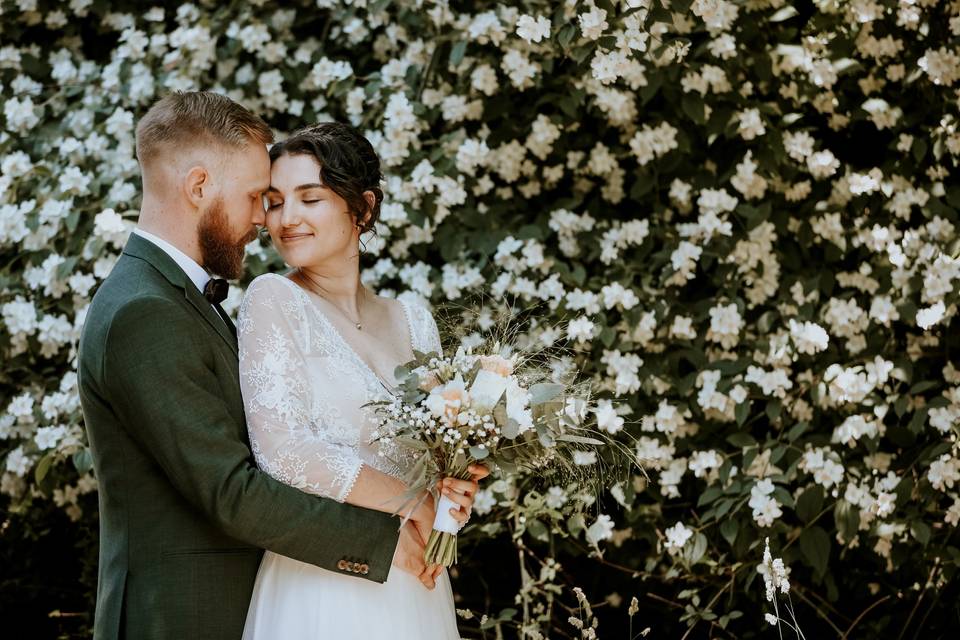 Jeune mariés près d'un mur de fleur, le marié avec un smocking et la mariée porte une robe en dentelle avec un bouquet de fleur dans les mains