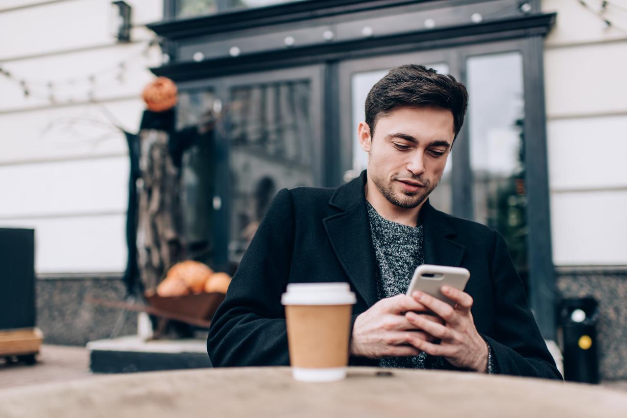 homme brun, jeune et élégant assis à la terrasse d'un café, téléphone en main et petit sourire de coin. il est en train de regarder ou lire quelque chose sur son écran. texto mignon pour homme, sms qui fait craquer un homme instantanément